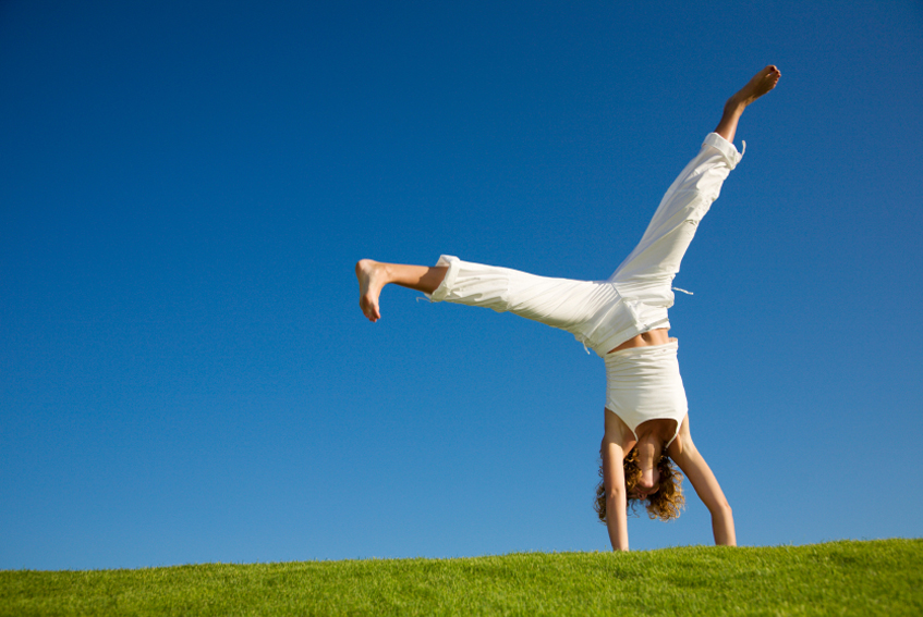 Woman wearing a white top and pants and doing a handstand on grass